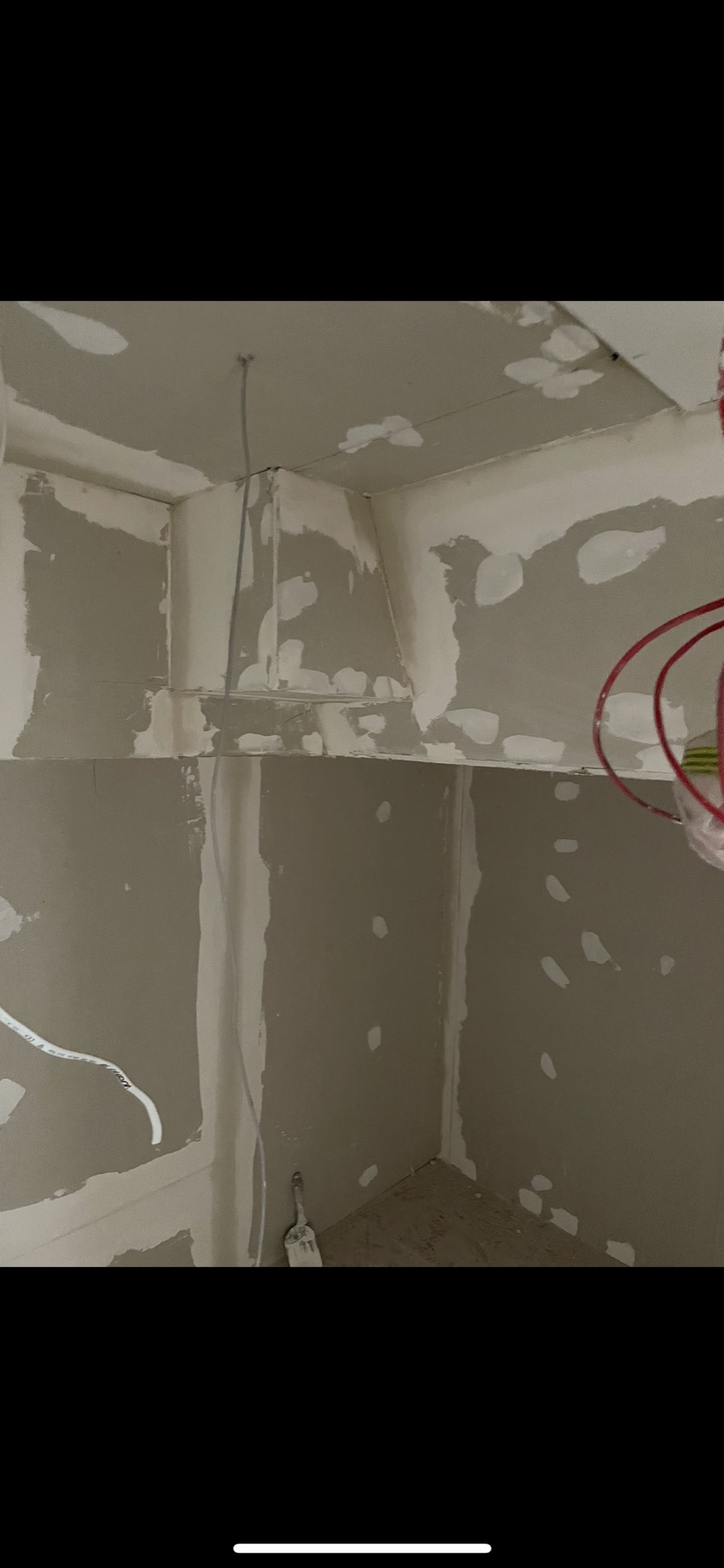 Niedokończone wnętrze z płyt kartonowo-gipsowych, widoczne łączenia szpachlowane, wiszące przewody elektryczne i pozostawiony pędzel. W tle fragment podłogi pokrytej zaprawą.