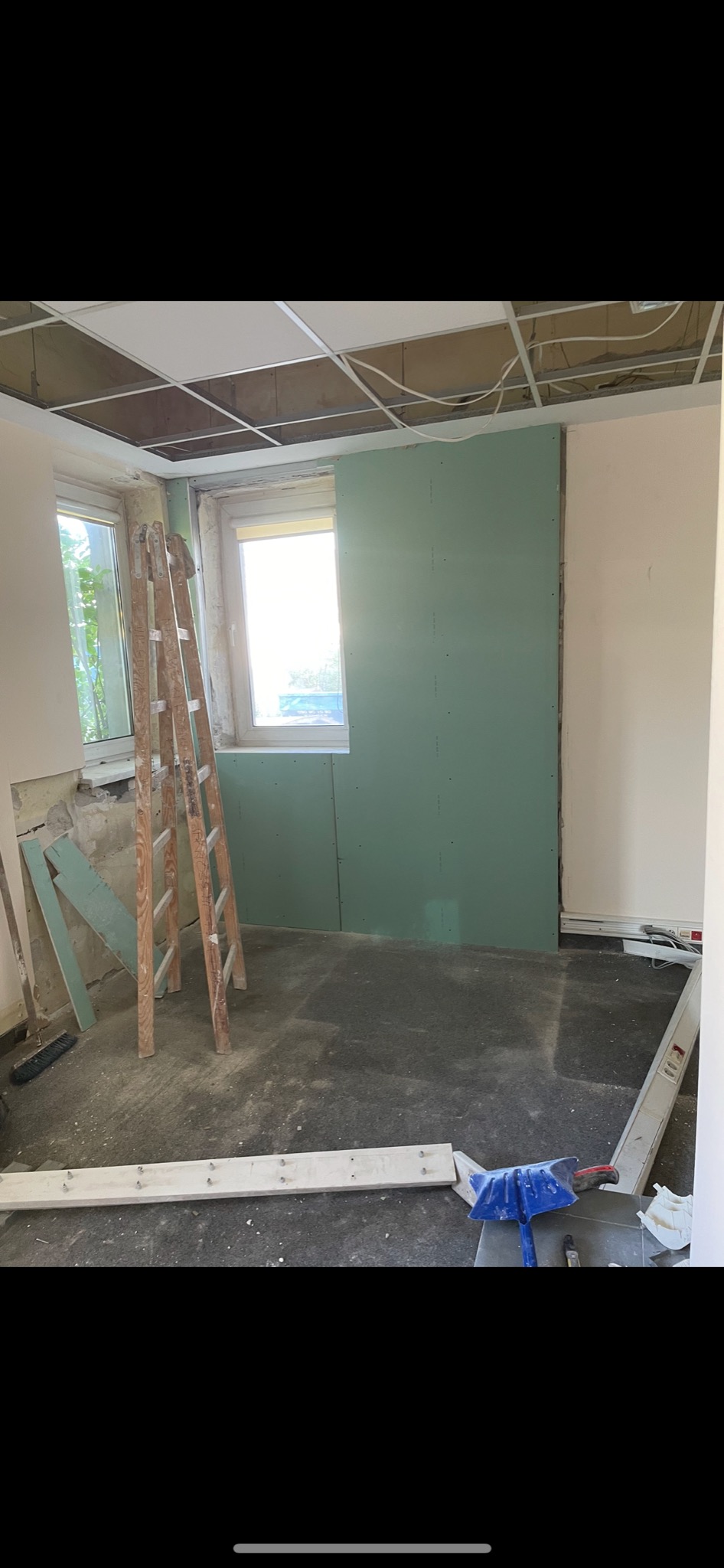 Pokój w trakcie remontu: zamontowane płyty gipsowo-kartonowe na ścianie, widoczny stelaż sufitu podwieszanego, drabina oparta o ścianę przy oknie, narzędzia i elementy konstrukcyjne na podłodze.
