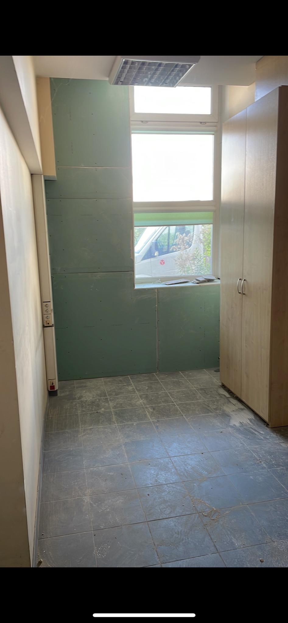 Wnętrze pomieszczenia w trakcie montażu płyt kartonowo-gipsowych na ścianach, widoczne szare płytki podłogowe, okno z widokiem na zewnątrz oraz szafa w rogu, instalacja elektryczna na ścianie.