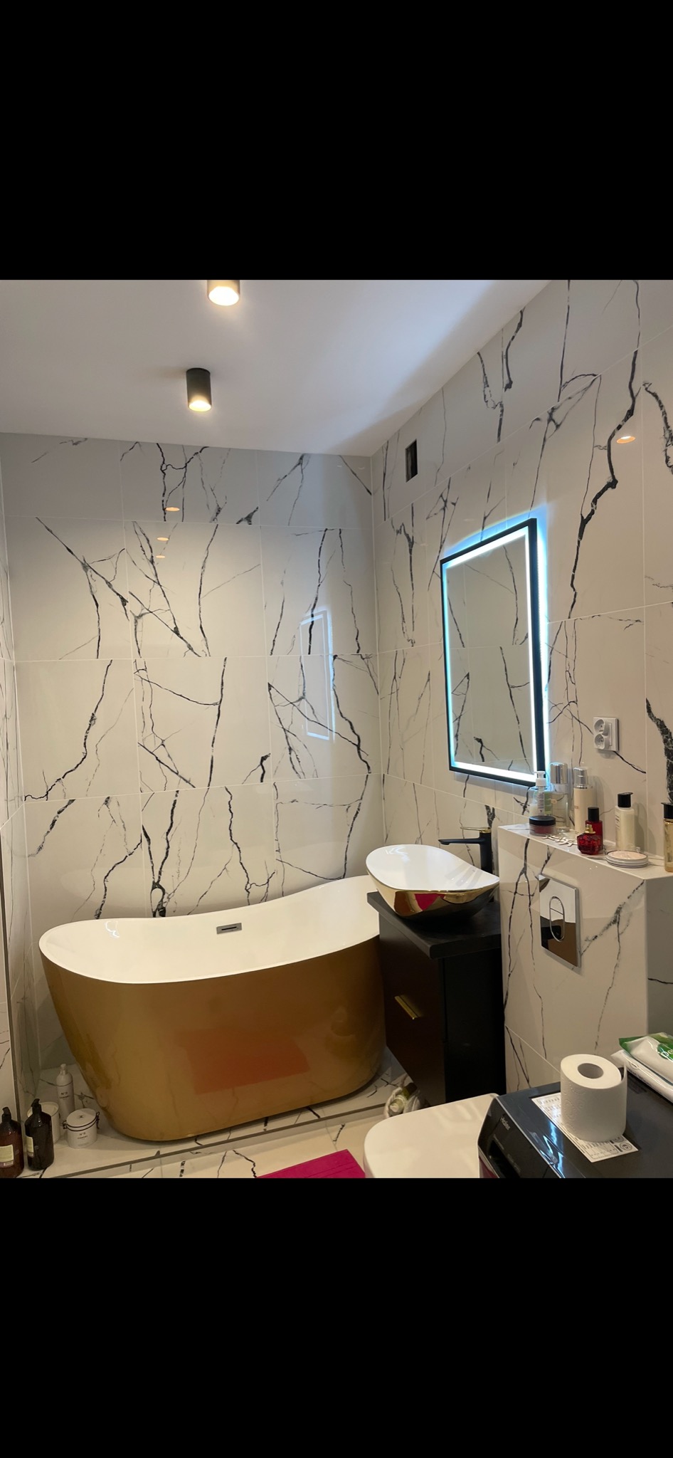 Nowoczesna łazienka z wolnostojącą, złotą wanną, umywalką nablatową i podświetlanym lustrem na tle białych płytek z czarnym marmurkowym wzorem.
