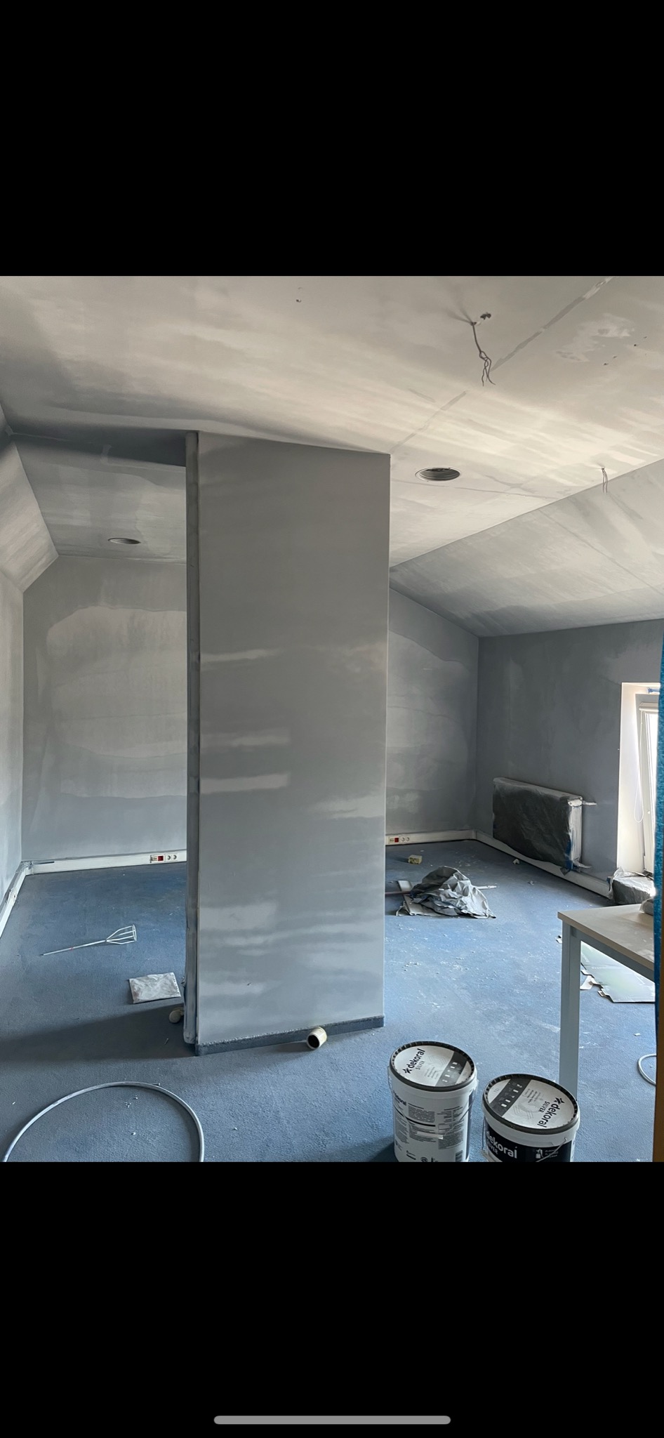 Wnętrze w trakcie remontu: ściany i sufit pokryte świeżo nałożoną gładzią, widoczne puszki farby Dekoral i narzędzia malarskie na podłodze zabezpieczonej folią.