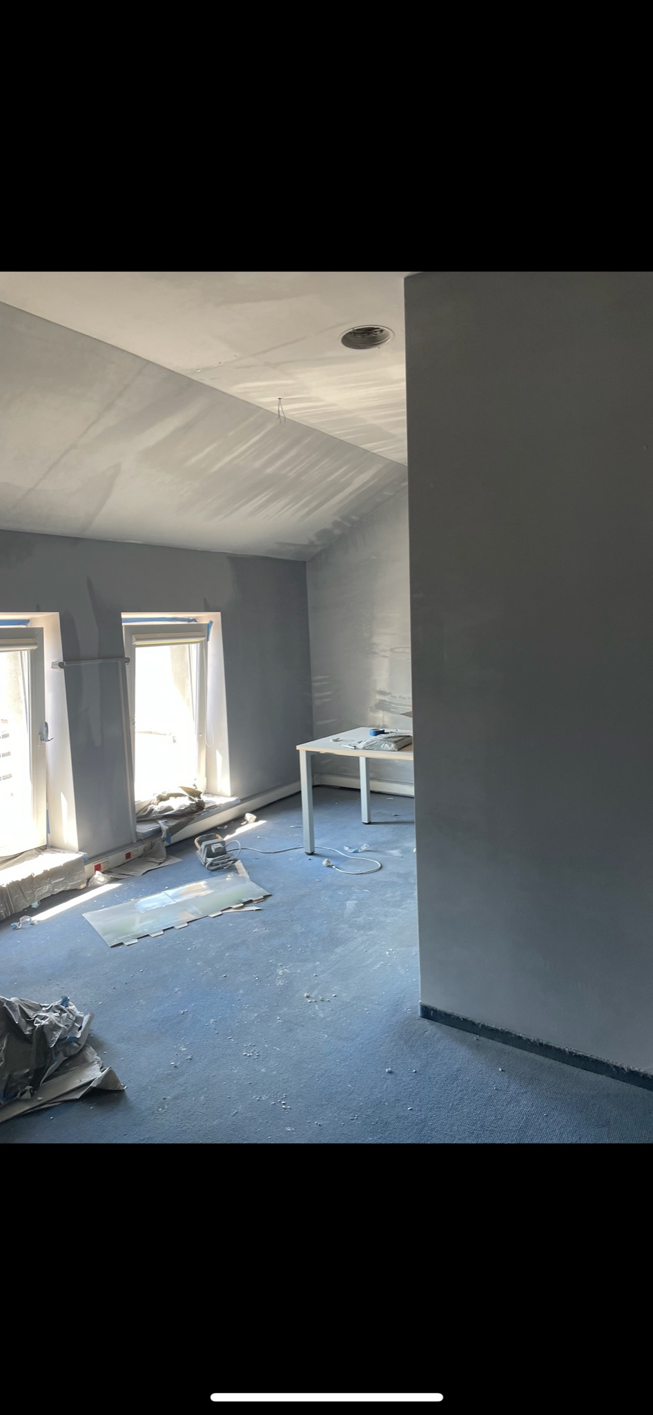 Pokój w trakcie malowania na szaro, widoczne narzędzia malarskie, zabezpieczone okna i niedokładności malowania na suficie