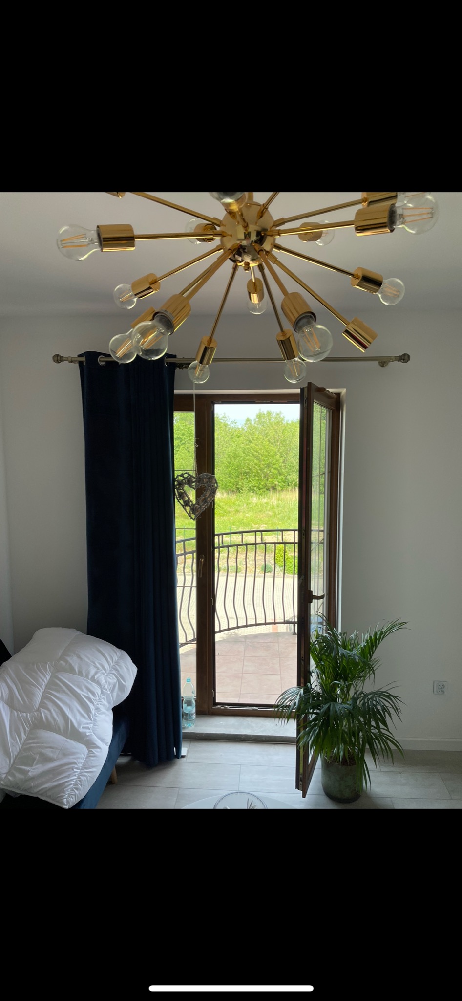 Jasne wnętrze z otwartymi drzwiami balkonowymi, przez które widać zieleń, z nowoczesną lampą sufitową w złotym kolorze, dekoracyjnym sercem na drzwiach i zieloną rośliną doniczkową.