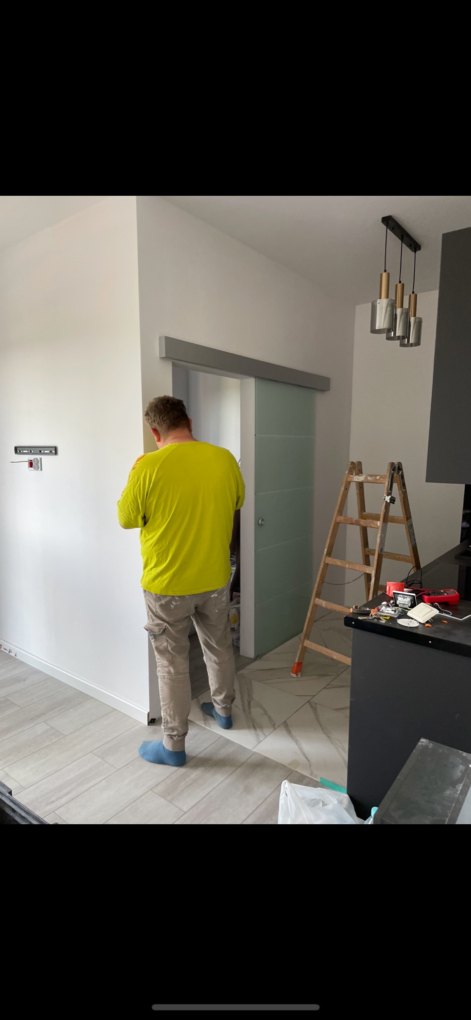 Pracownik w jaskrawej koszulce montuje listwę przypodłogową przy szklanych drzwiach przesuwnych w wyremontowanym wnętrzu, widoczna drabina i narzędzia na blacie.