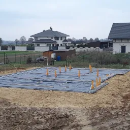 Przygotowany teren pod fundamenty domu jednorodzinnego, widoczna czarna folia izolacyjna, pionowe rury kanalizacyjne i drewniane listwy na piasku, w tle zabudowa jednorodzinna i pochmurne niebo.