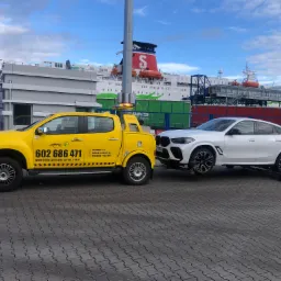Żółty samochód pomocy drogowej z numerem telefonu na tle promu pasażerskiego holuje białe BMW X6 na wózkach transportowych po brukowanym placu.