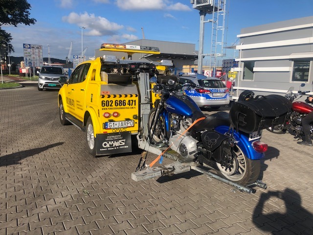 Żółty samochód pomocy drogowej z załadowanym niebieskim motocyklem Harley Davidson na tle parkingu z kostki brukowej i radiowozu policyjnego.