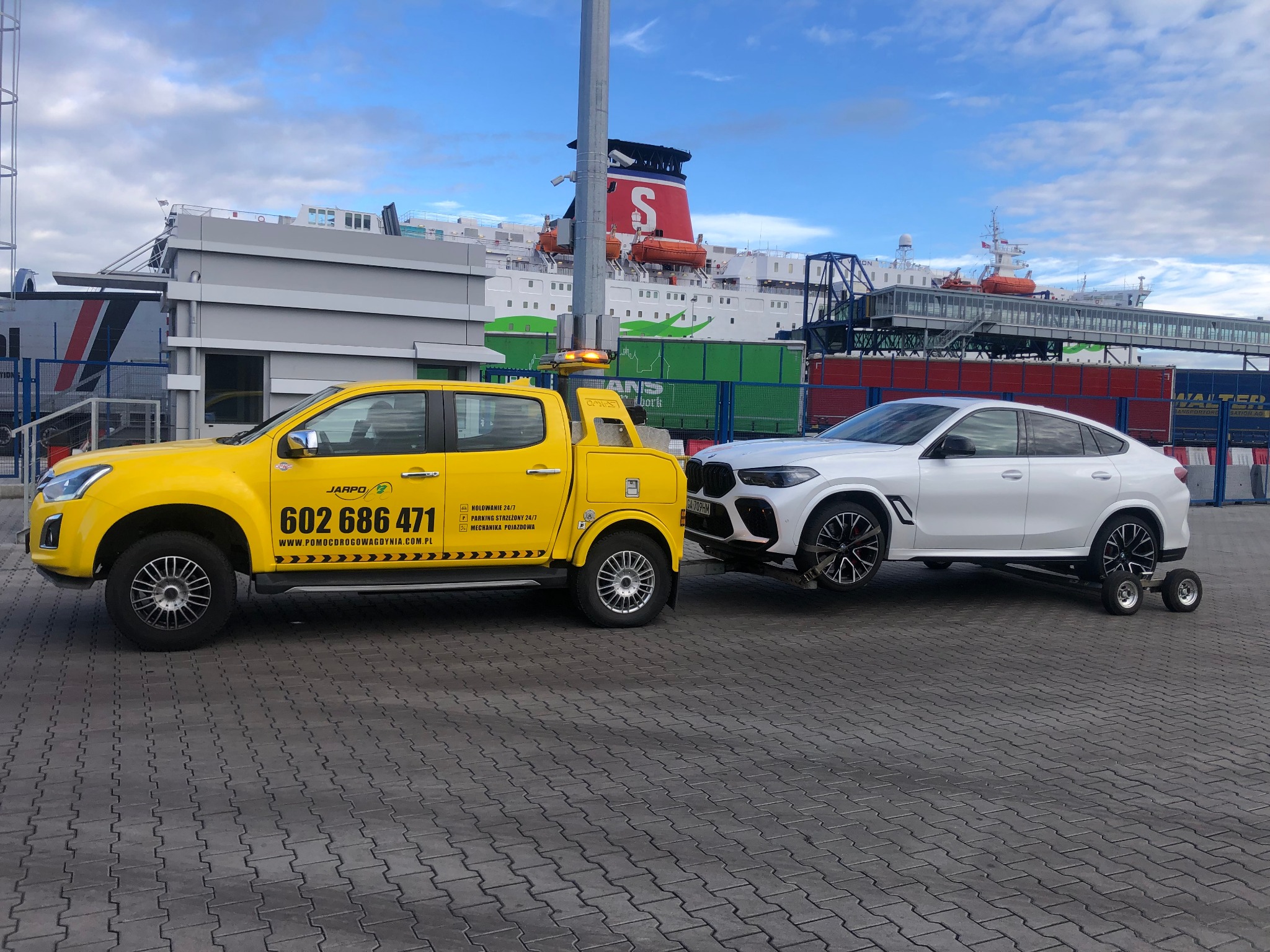 Żółty samochód pomocy drogowej z numerem telefonu na tle promu pasażerskiego holuje białe BMW X6 na wózkach transportowych po brukowanym placu.