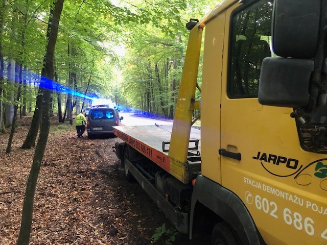 Żółty samochód pomocy drogowej z lawetą na poboczu leśnej drogi, załadowywany jest szary samochód osobowy, w tle widoczne niebieskie światła