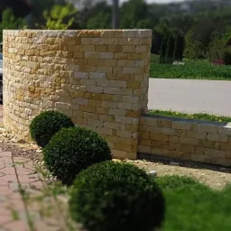 Zakrzywiony murek z piaskowca, zakończony prostokątnym słupkiem, z trzema kulistymi krzewami na pierwszym planie, droga w tle.