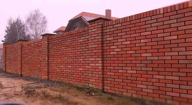 Długi, ceglany mur ogrodzeniowy z filarami zwieńczonymi czapkami, wykonany z czerwonej cegły klinkierowej z ciemną fugą, widok na dom z kominem w tle.