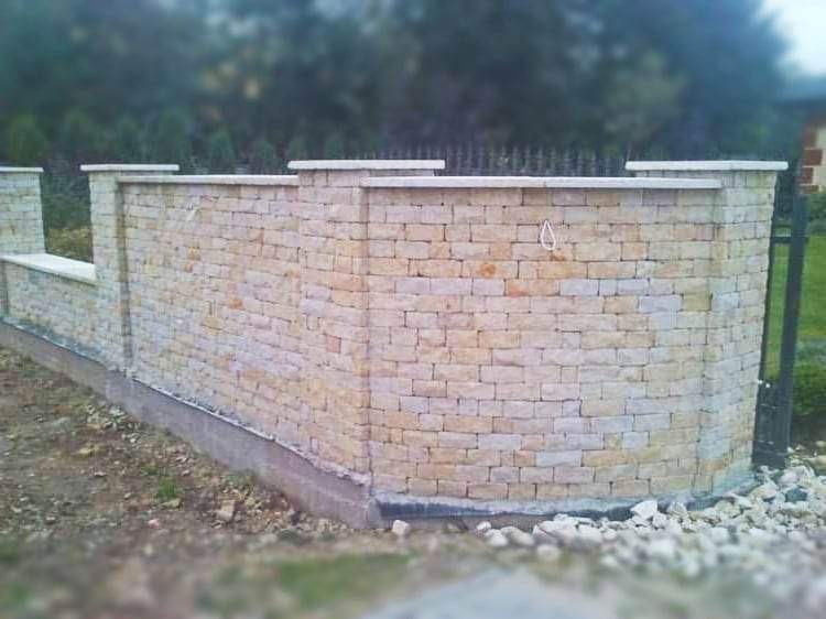 Murek ogrodzeniowy z piaskowca w kształcie łuku, zwieńczony płaskimi elementami, z fragmentem metalowej bramy w tle.