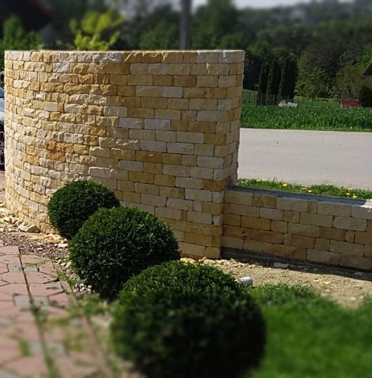 Zakrzywiony murek z piaskowca, zakończony prostokątnym słupkiem, z trzema kulistymi krzewami na pierwszym planie, droga w tle.