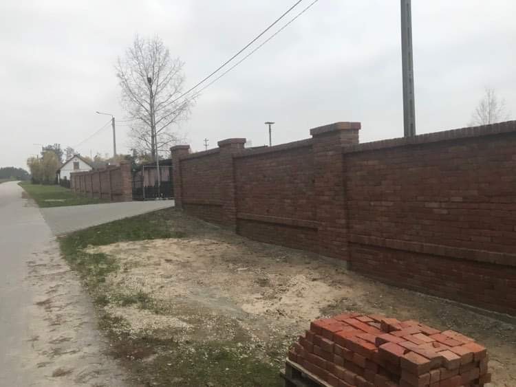 Ceglany mur ogrodzeniowy z filarami, widok z zewnątrz, sterta cegieł na palecie w pierwszym planie, szara nawierzchnia drogi po lewej stronie, drzewo bez liści w tle.