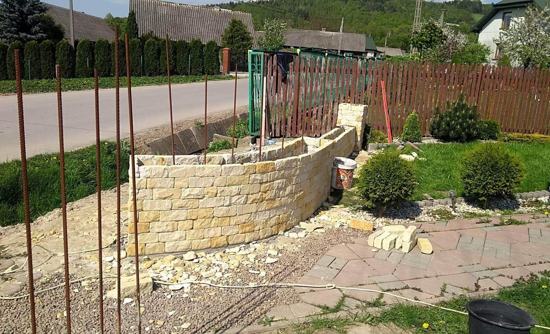 Budowa niskiego, zakrzywionego murku z piaskowca w ogrodzie, widoczne wystające pręty zbrojeniowe, częściowo ułożona kostka brukowa i wiaderko po zaprawie.