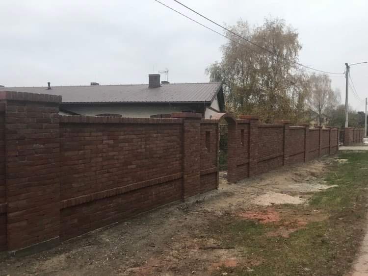 Ceglany mur ogrodzeniowy z furtką zakończoną łukiem, widok z zewnątrz na tle szarego nieba i drzew bez liści.