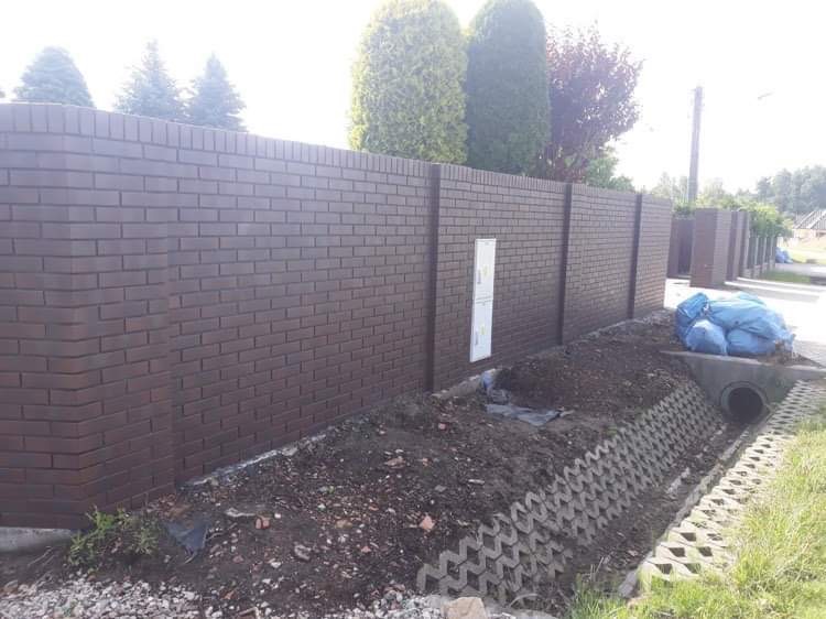 Nowoczesne, brązowe ogrodzenie z cegły klinkierowej z betonowymi słupkami, częściowo obsypane ziemią, z widoczną skrzynką elektryczną na jednym z przęseł.
