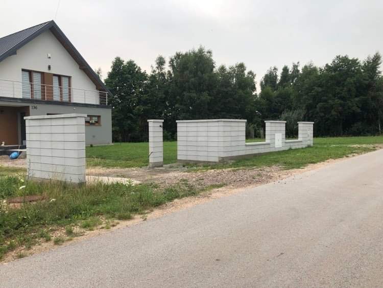 Wznoszone ogrodzenie z szarych, betonowych bloczków w surowym stanie, widoczne słupki i fragment podmurówki, na tle domu jednorodzinnego i zieleni.