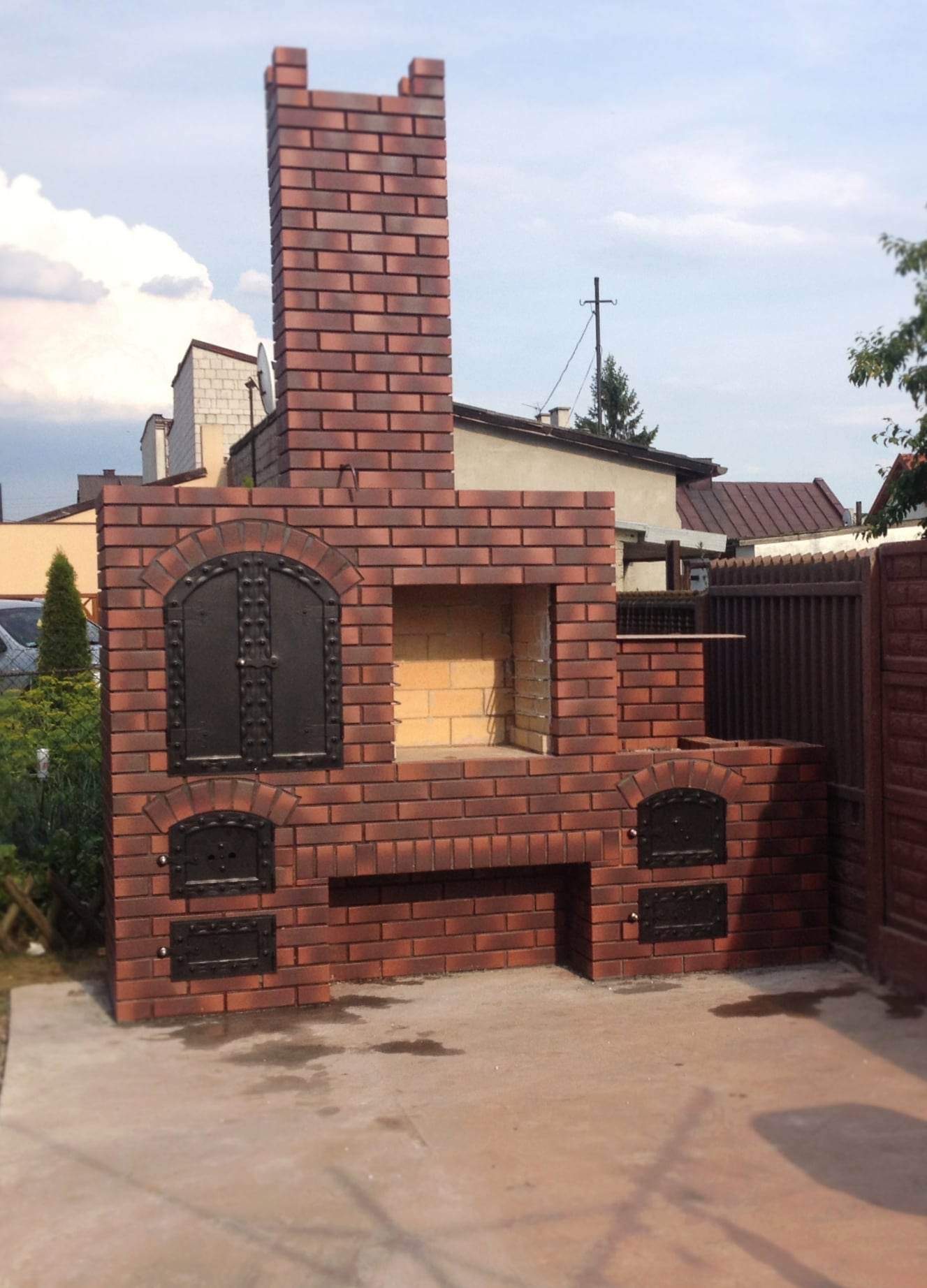 Murowany grill ogrodowy z czerwonej cegły z czarnymi, kutymi drzwiczkami, umieszczony na betonowym podłożu, z widocznym fragmentem ogrodzenia w tle.