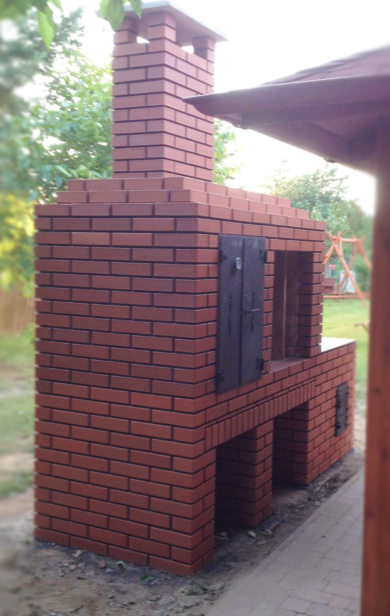 Murowany grill ogrodowy z czerwonej cegły z czarnymi drzwiczkami, kominem i zadaszeniem, otoczony zielenią.