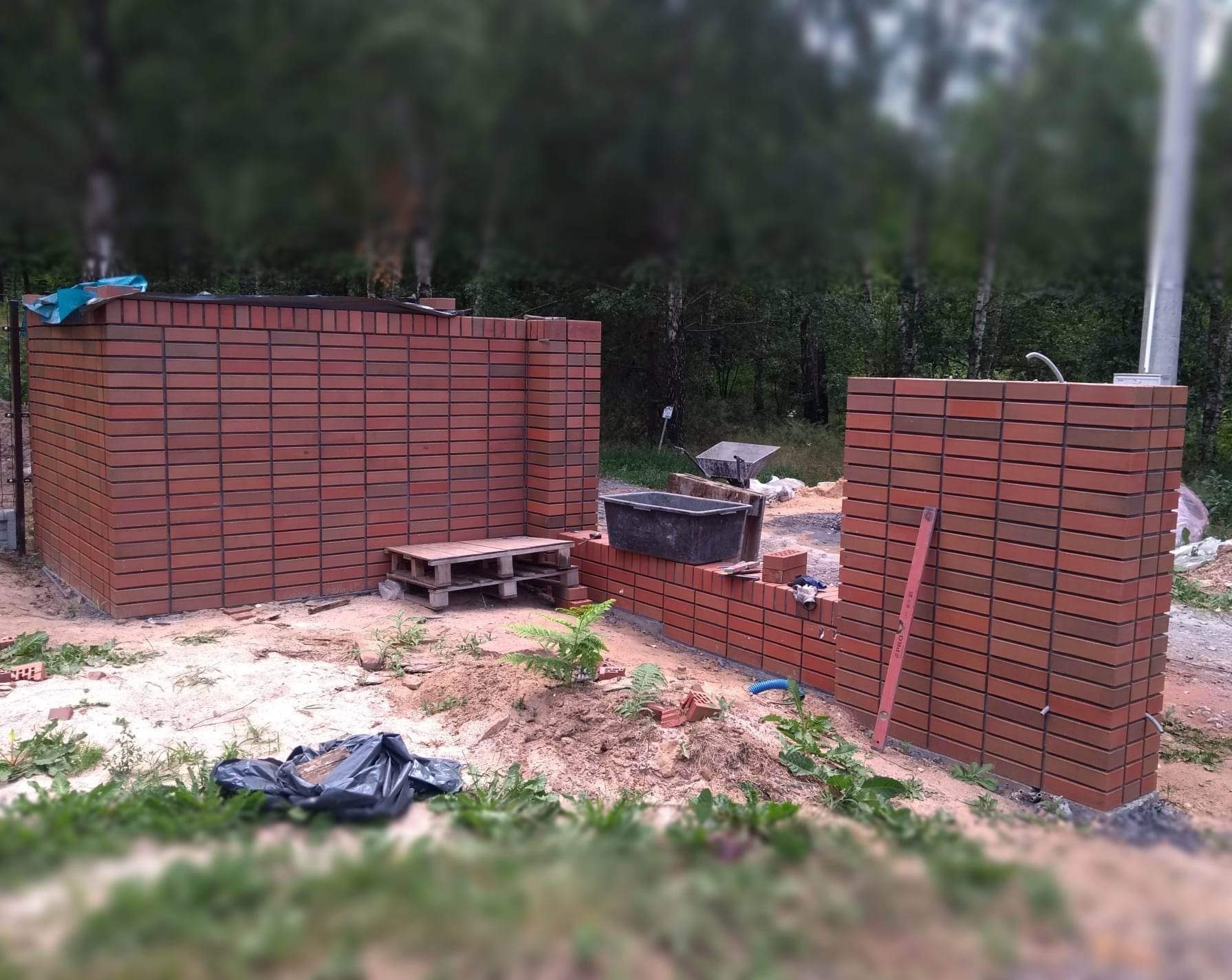 Wznoszenie ogrodzenia z czerwonej cegły klinkierowej w trakcie budowy, widoczne narzędzia i materiały budowlane, tło zalesione.