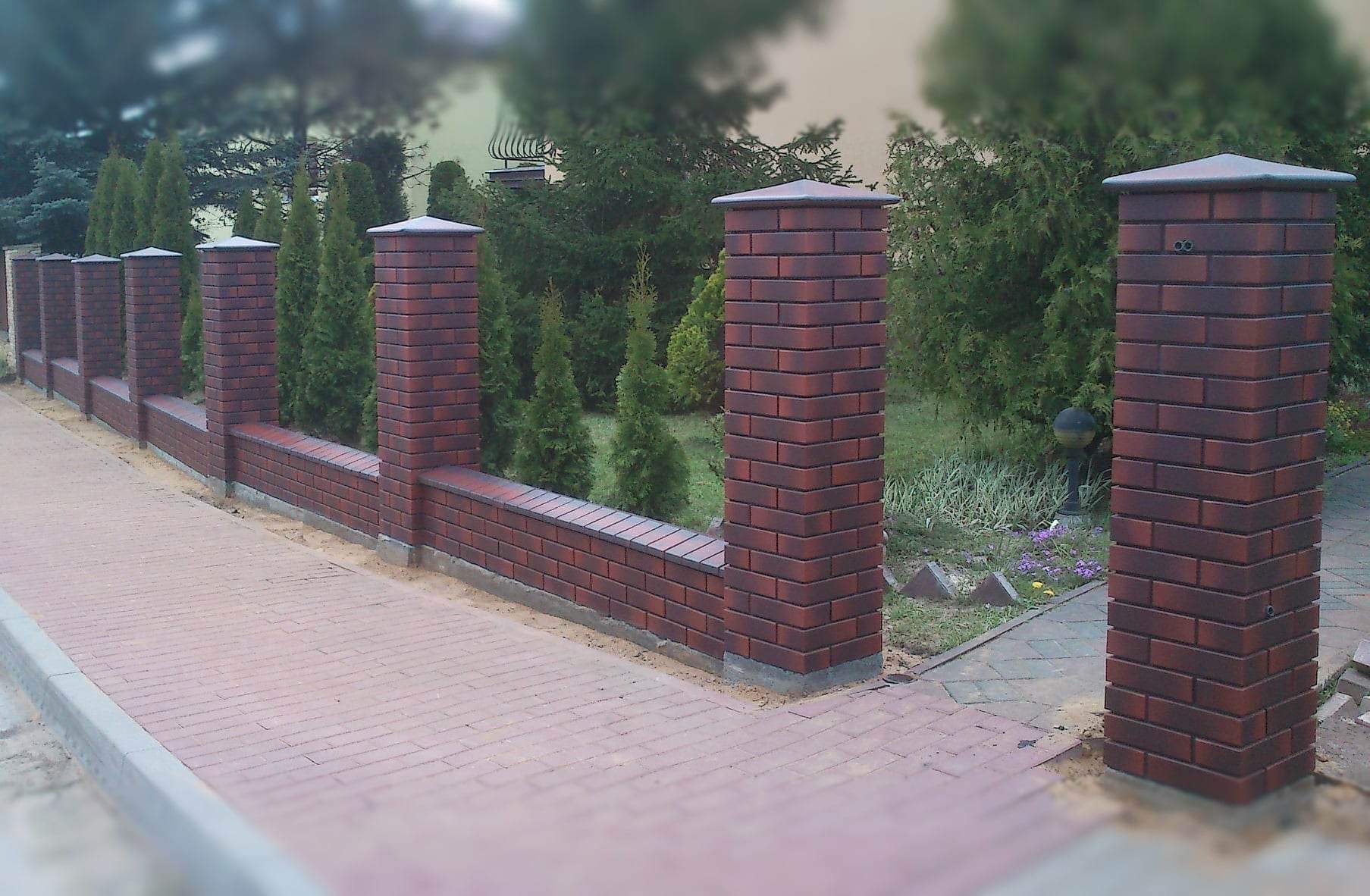 Ceglany mur z filarami i ciemnymi czapkami, oddzielający posesję od chodnika, widok z perspektywy pieszego, zieleń w tle.