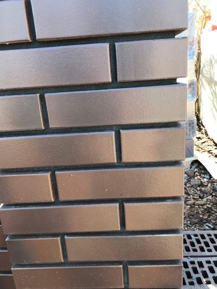 Fragment brązowego ogrodzenia z cegieł klinkierowych, widoczna struktura i faktura materiału, z elementami kanalizacji liniowej w tle.