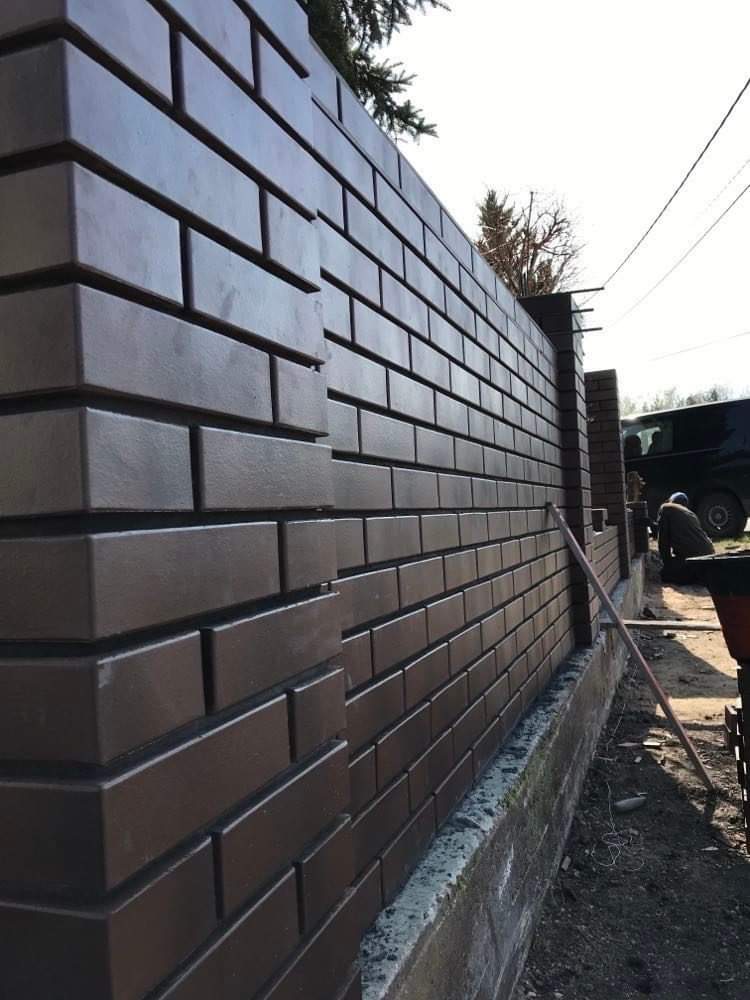 Fragment brązowego ogrodzenia z cegły klinkierowej na betonowej podmurówce, widoczny pracownik w tle i fragment pojazdu.
