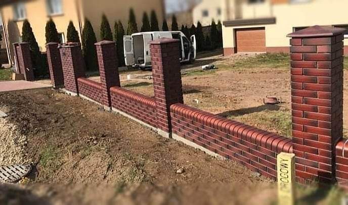 Nowo wybudowany, niski murek z cegły klinkierowej z filarami, oddzielający posesję od chodnika; w tle biały bus z otwartymi drzwiami.