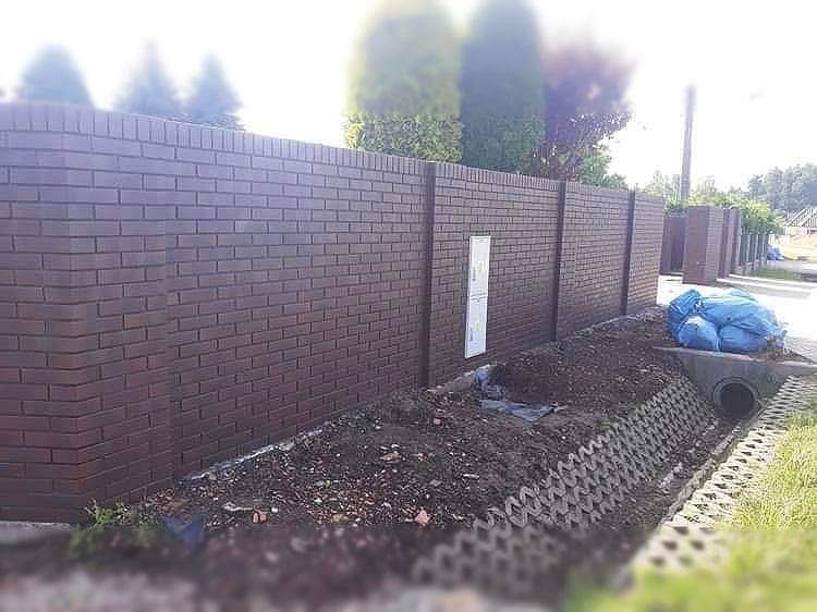 Nowoczesny, brązowy mur z cegły klinkierowej, pełniący funkcję ogrodzenia, z widoczną skrzynką elektryczną wkomponowaną w konstrukcję, obok świeżo usypana ziemia i niebieski worek.