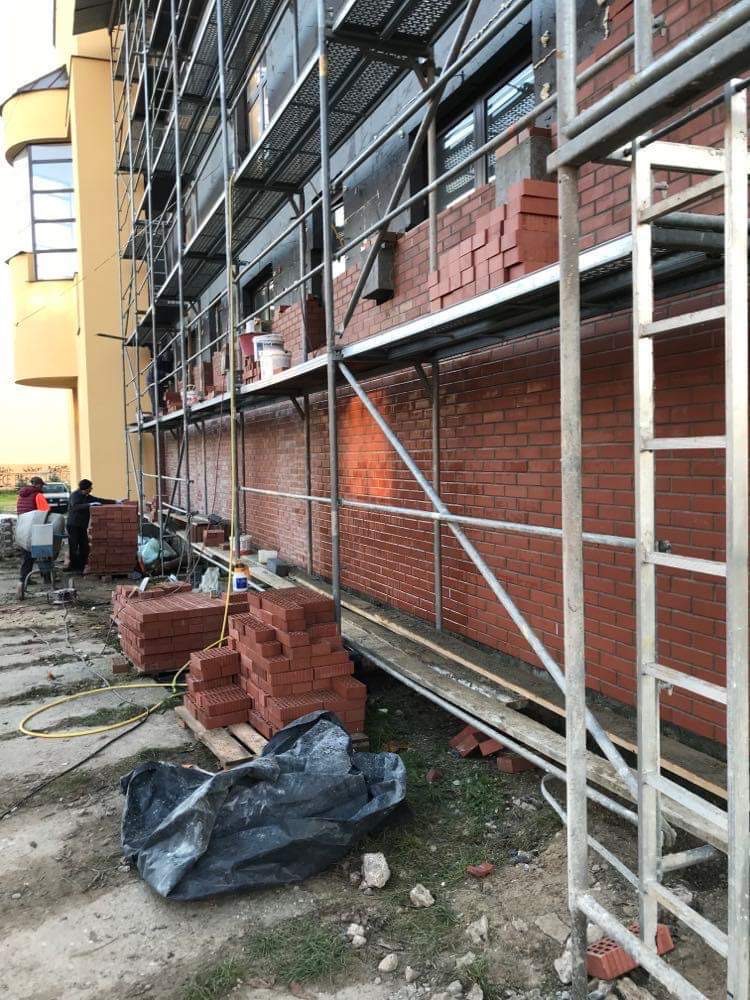 Budowa ogrodzenia z czerwonej cegły, widoczne rusztowanie i materiały budowlane, kilku pracowników w oddali.
