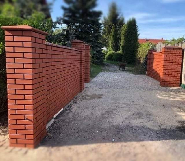 Czerwony, ceglany mur ogrodzeniowy z filarami, prowadzący do żwirowej ścieżki w ogrodzie.