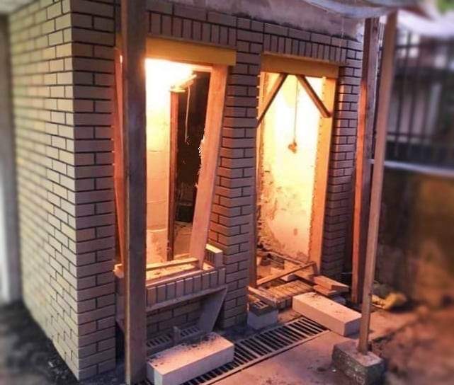 Wznoszenie murowanej konstrukcji, prawdopodobnie bramy lub ogrodzenia, z widocznymi drewnianymi elementami wspierającymi i kratką odpływową w podstawie.