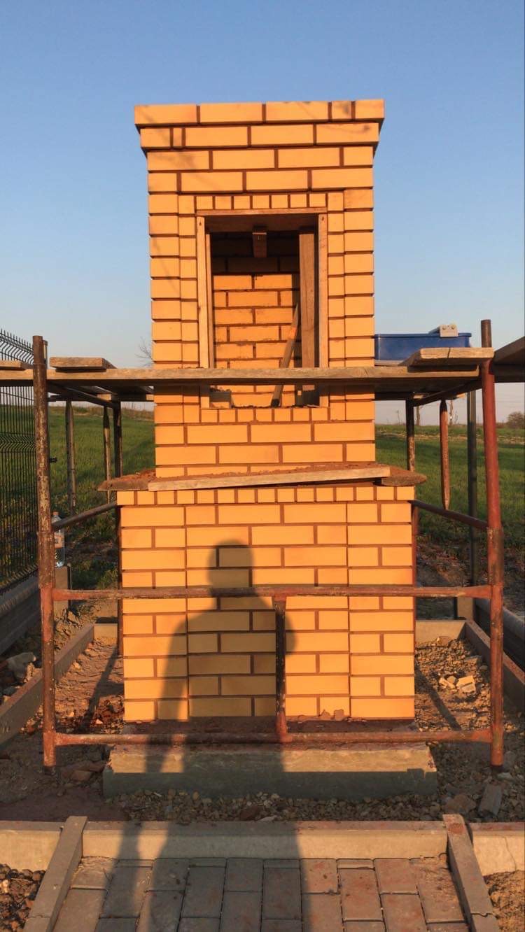 Wznoszona z żółtej cegły konstrukcja z otworem, prawdopodobnie pod grilla lub wędzarnię, otoczona rusztowaniem i z widocznym cieniem osoby fotografującej.