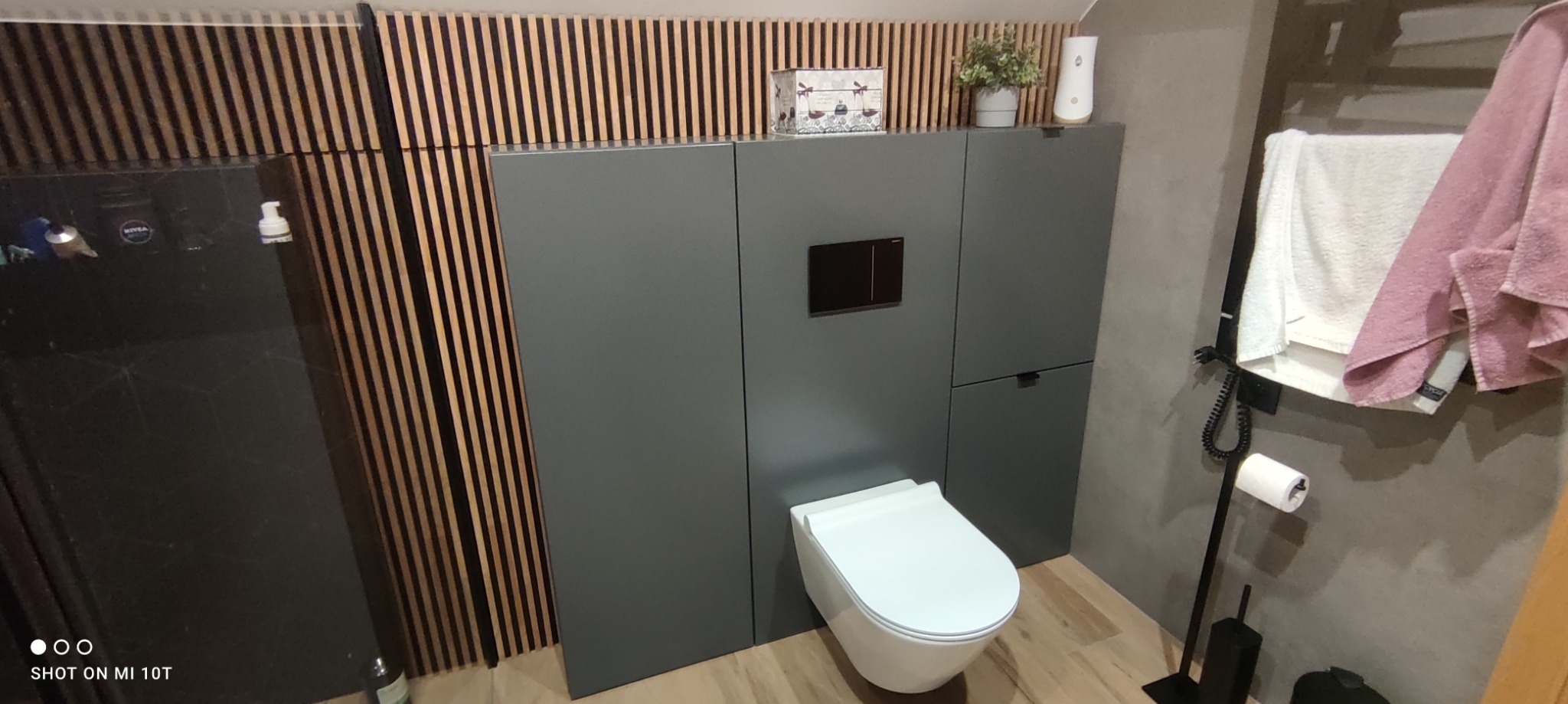 Nowoczesna toaleta podwieszana z zabudową w kolorze szarym, dopasowaną do drewnianej boazerii ściennej. Na szafkach dekoracje i dozownik, obok grzejnik drabinkowy z ręcznikami.