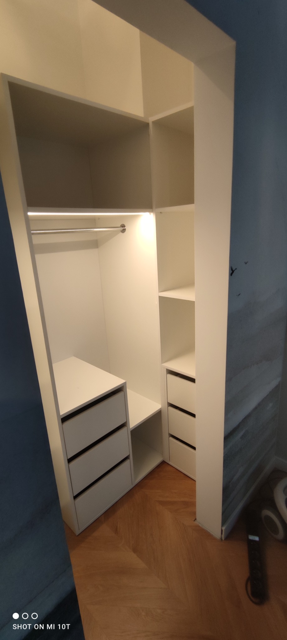 Wnętrze białej szafy wnękowej z szufladami i drążkiem, oświetlone od góry, na parkiecie ułożonym w jodełkę, z fragmentem ściany z motywem górskim w tle.