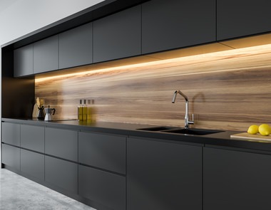 Nowoczesna kuchnia z czarnymi, matowymi szafkami i drewnianym panelem ściennym podświetlonym listwą LED. Na blacie widoczny czarny zlew, bateria, ekspres do kawy, butelki z olejem i deska z cytrynami.