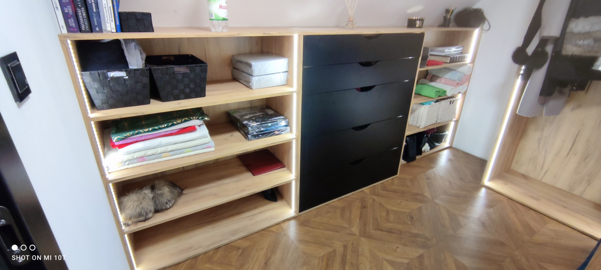 Półki z jasnego drewna z podświetleniem LED i czarnymi szufladami, wypełnione ubraniami, pudełkami i dekoracjami w pomieszczeniu ze skośnym sufitem.