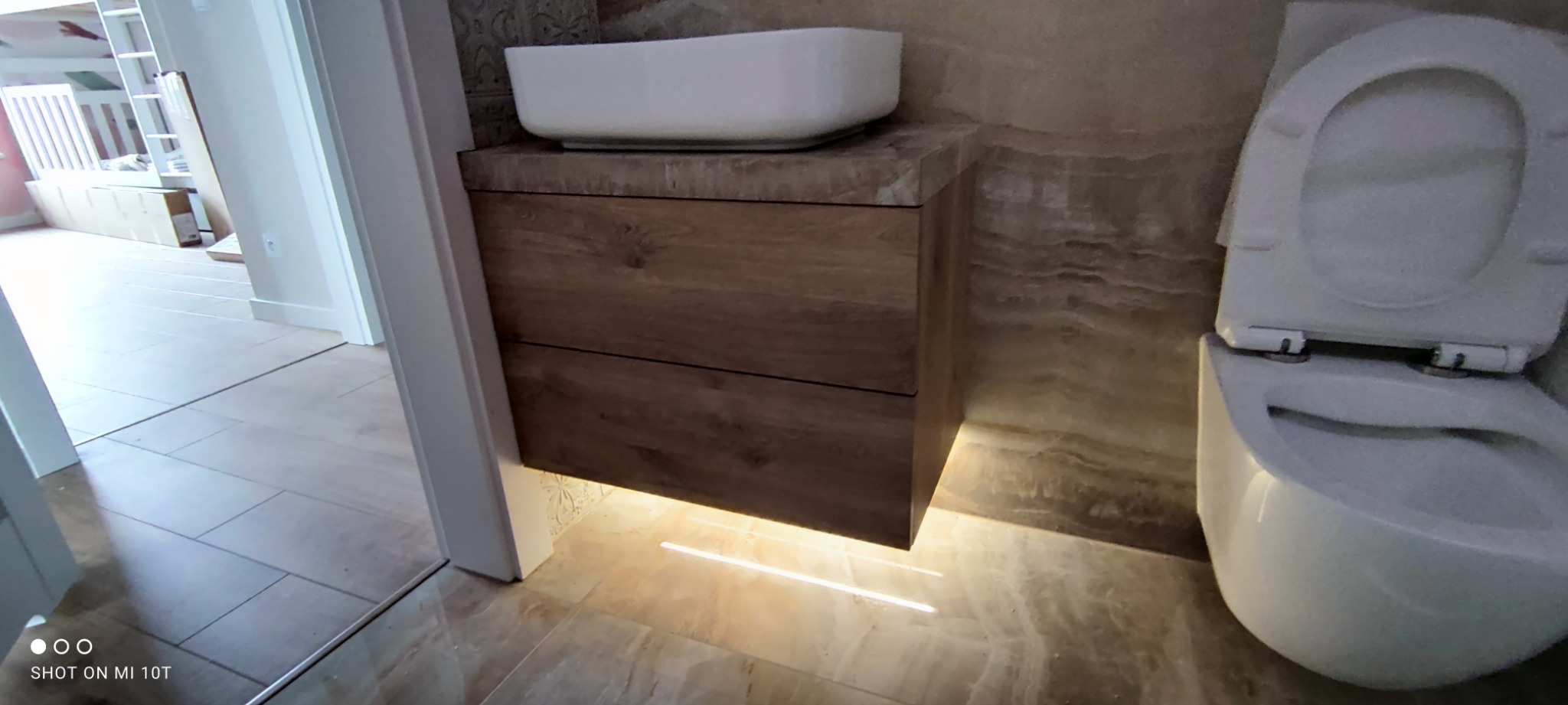 Nowoczesna łazienka z podświetlaną szafką pod umywalkę z drewnianym frontem i białą umywalką nablatową, obok podwieszana toaleta.