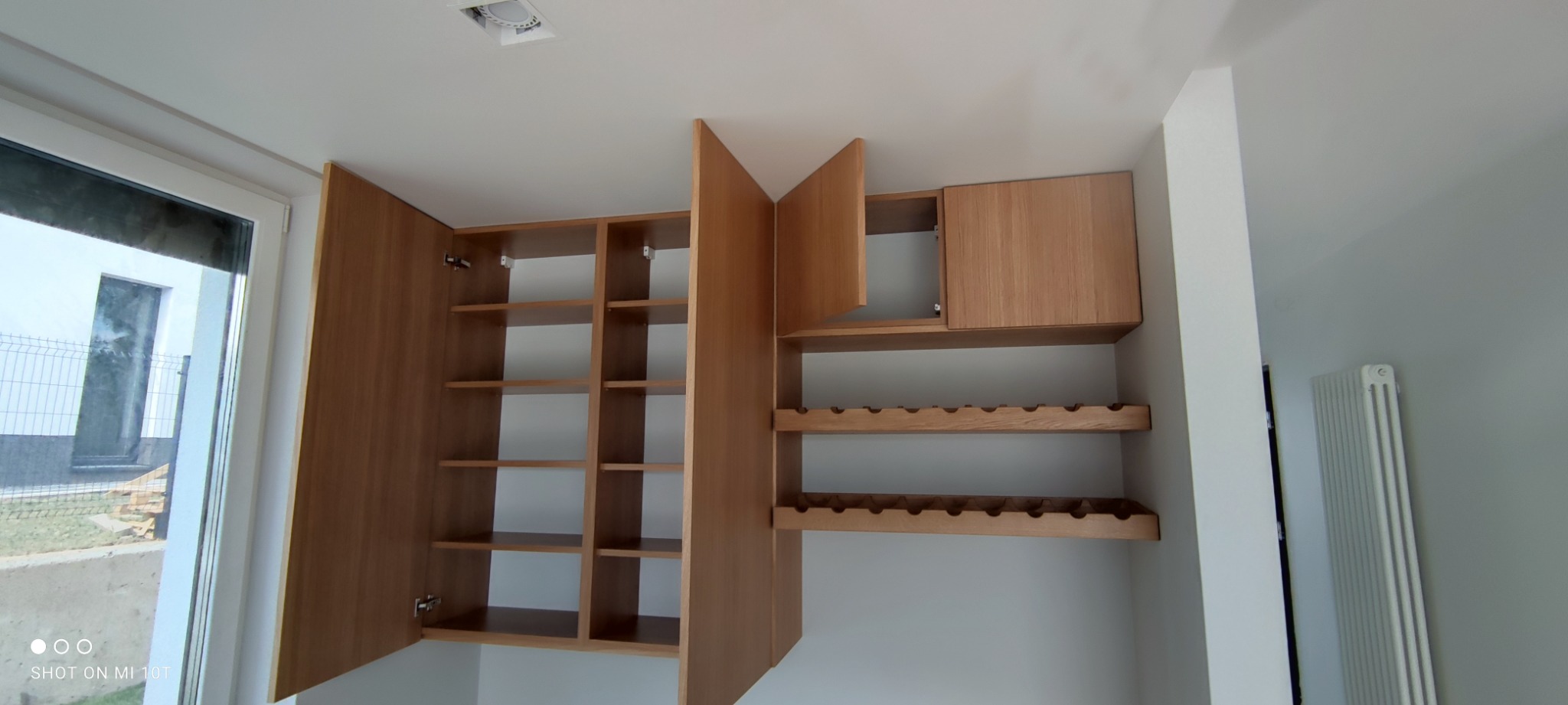 Drewniana zabudowa meblowa z otwartymi półkami i półkami na wino, widoczna z bliska, z oknem po lewej stronie.