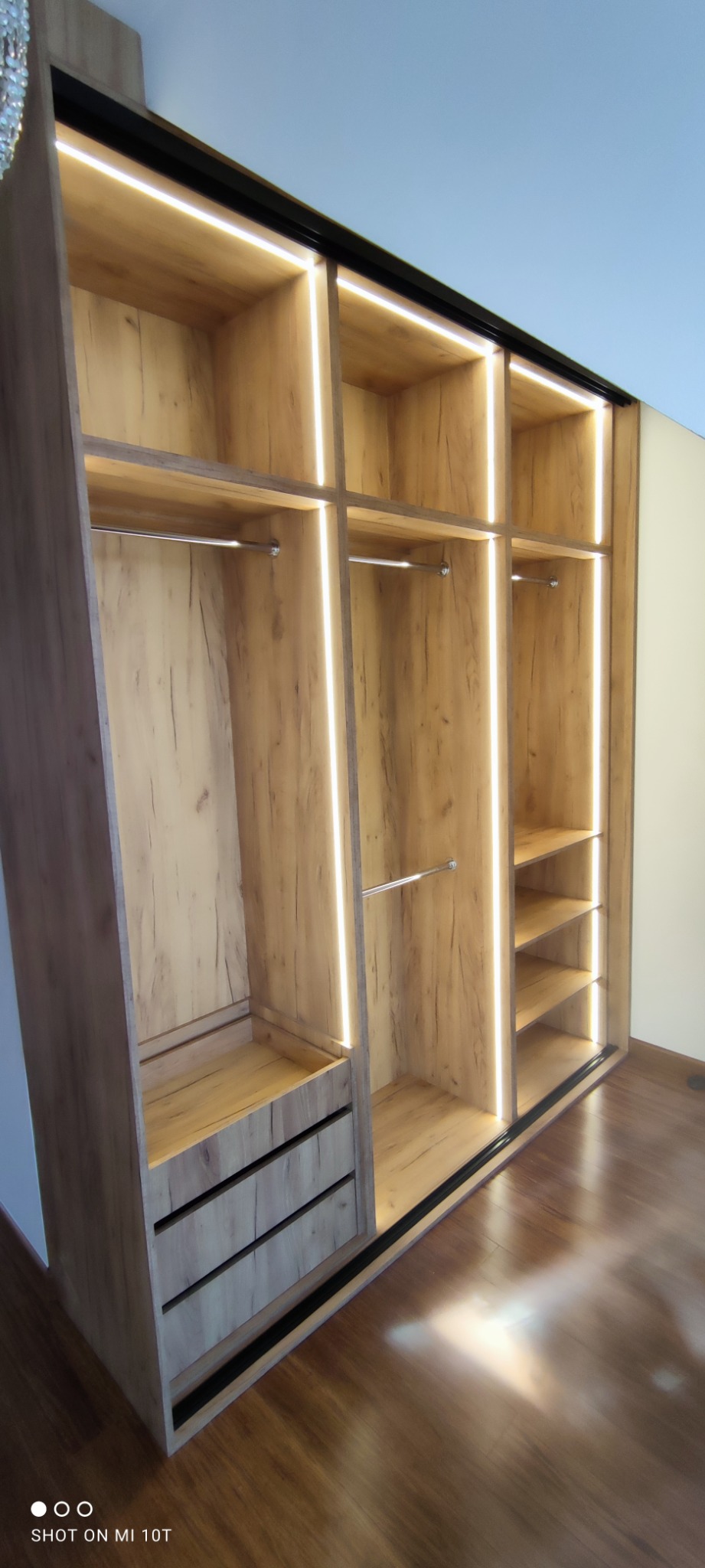 Wnętrze szafy wnękowej z przesuwnymi drzwiami, oświetleniem LED i drewnianym wykończeniem, widoczne półki, drążki na ubrania oraz szuflady.