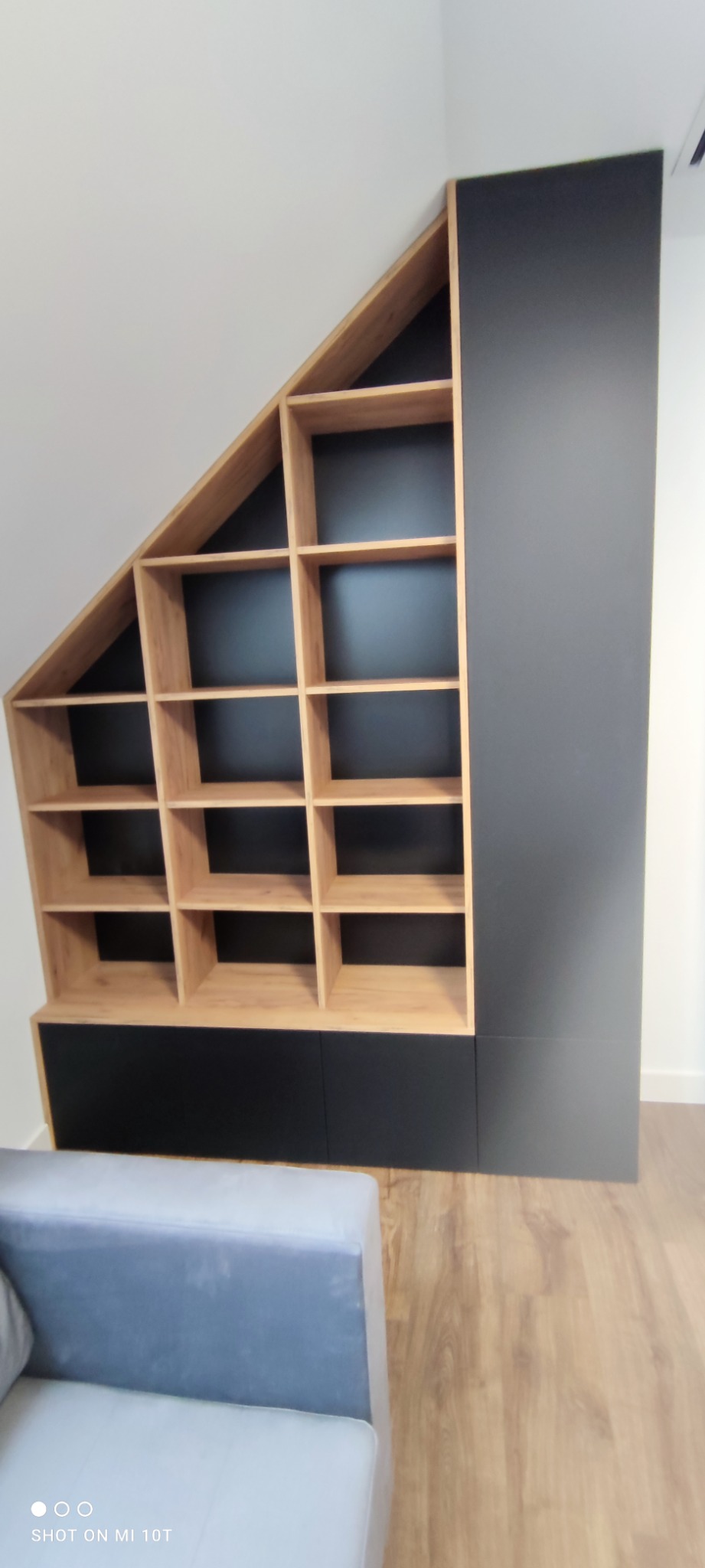 Szafa wnękowa z drewnianymi półkami i czarnymi frontami, dopasowana do skosu dachu, widoczny fragment szarej sofy.