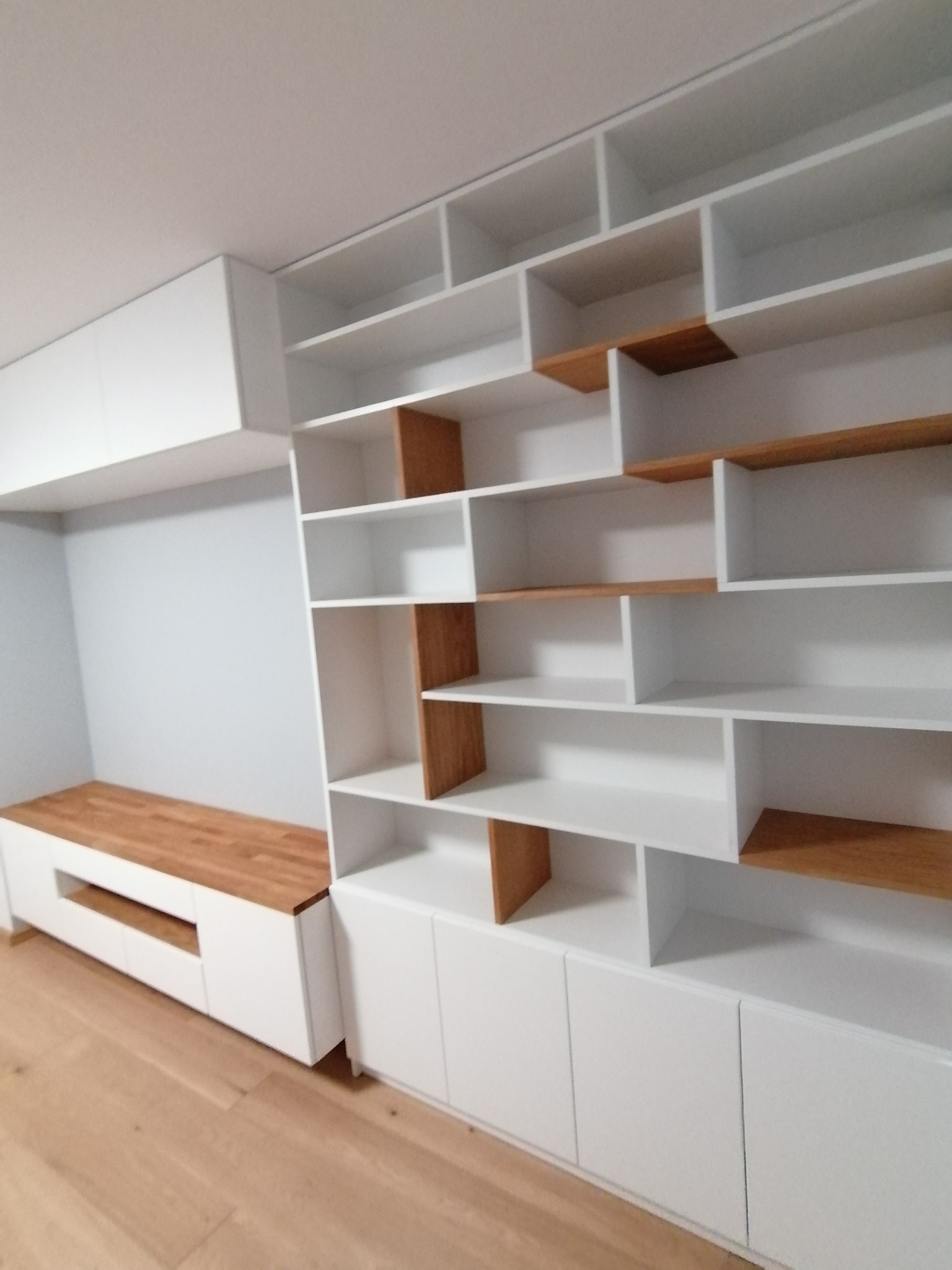 Biała zabudowa meblowa z drewnianymi akcentami, obejmująca szafki dolne i półki otwarte, tworząca spójną całość z drewnianym blatem siedziska.
