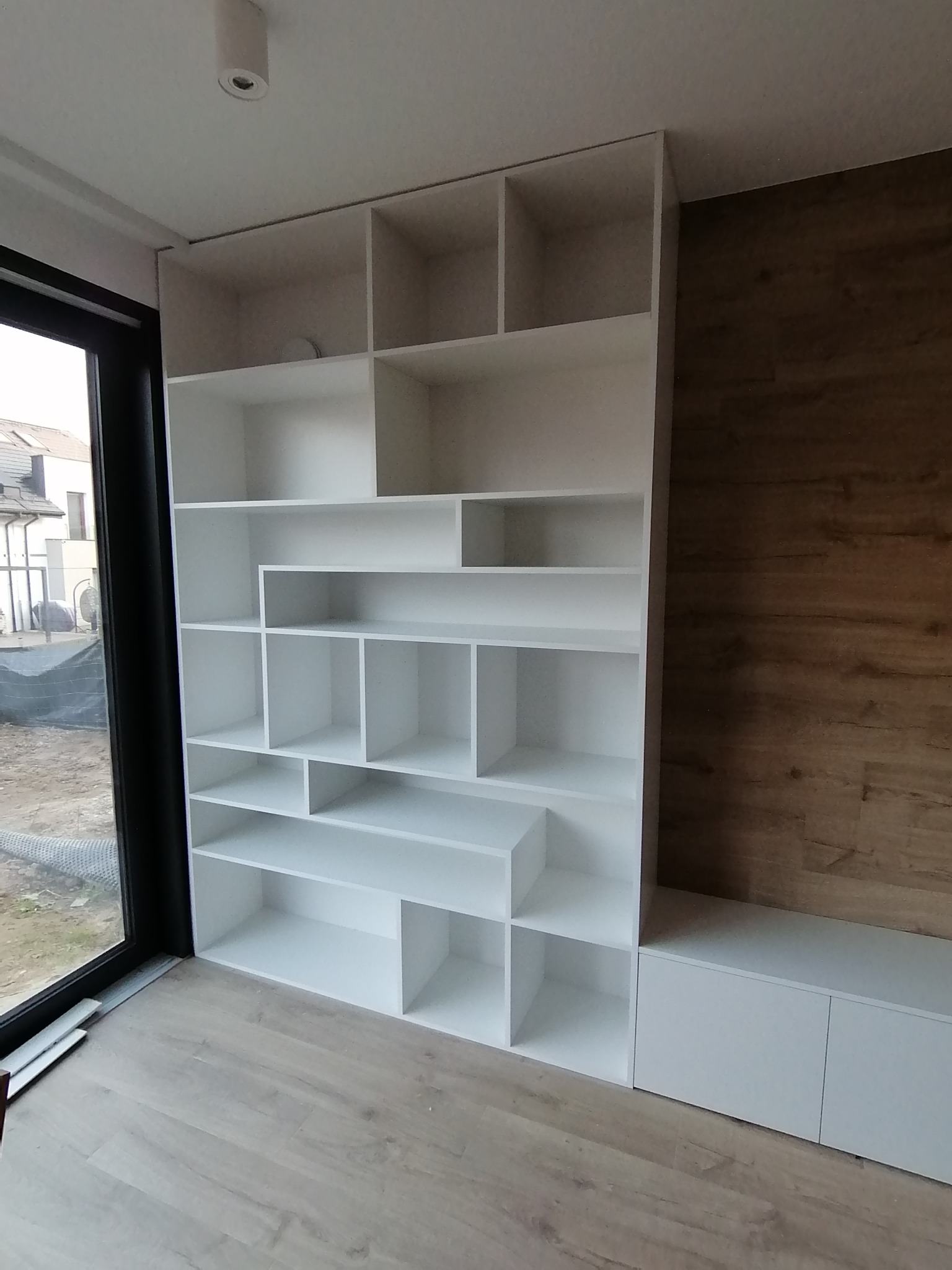 Biała, asymetryczna zabudowa meblowa z półkami, zintegrowana z niską szafką RTV, na tle ściany wykończonej panelami drewnopodobnymi, widoczna przez okno budowa na zewnątrz.