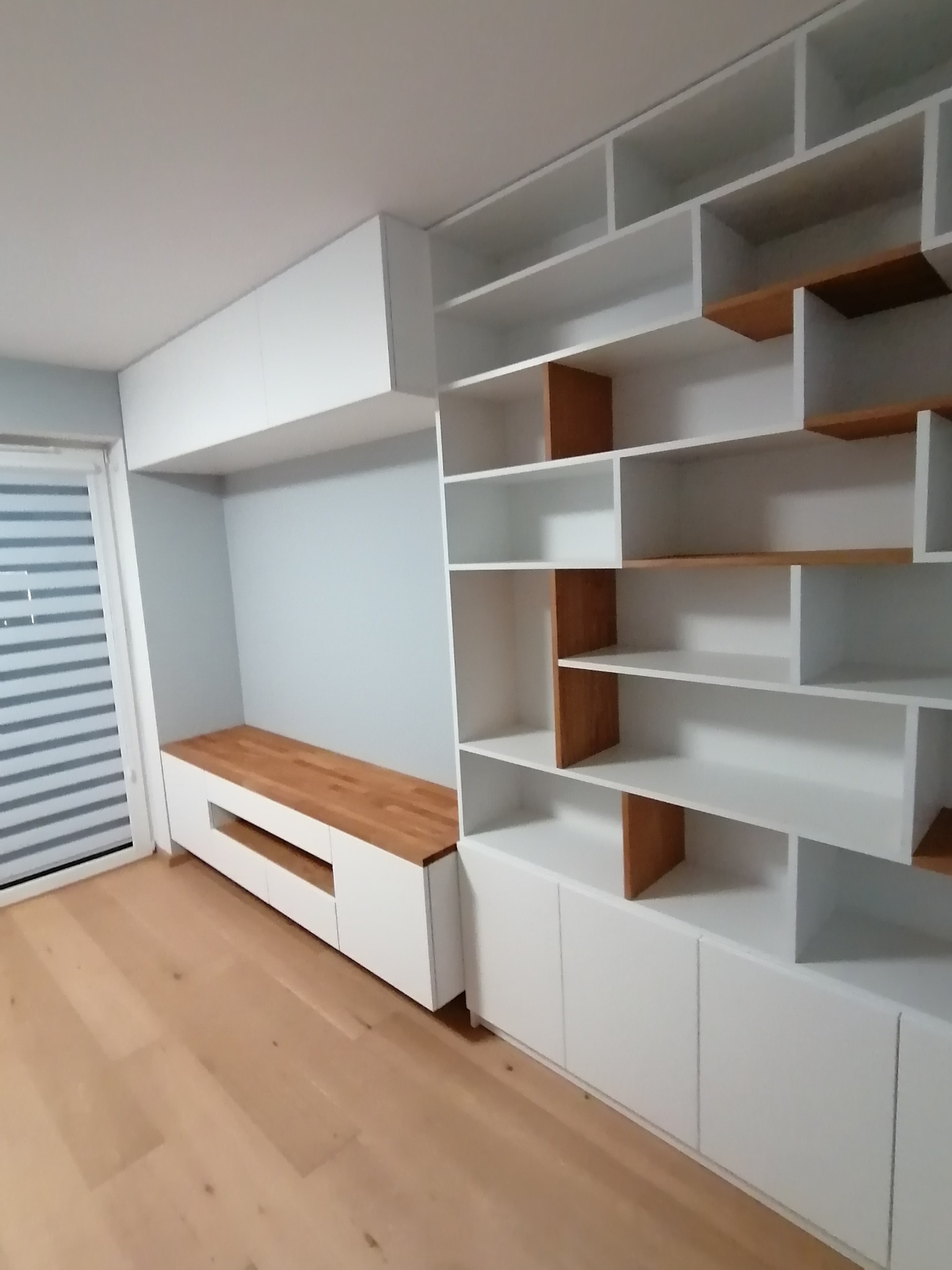 Biała zabudowa meblowa z drewnianymi akcentami, obejmująca regał z asymetrycznymi półkami i szafkę RTV z drewnianym blatem, na tle jasnej podłogi i okna z żaluzjami.