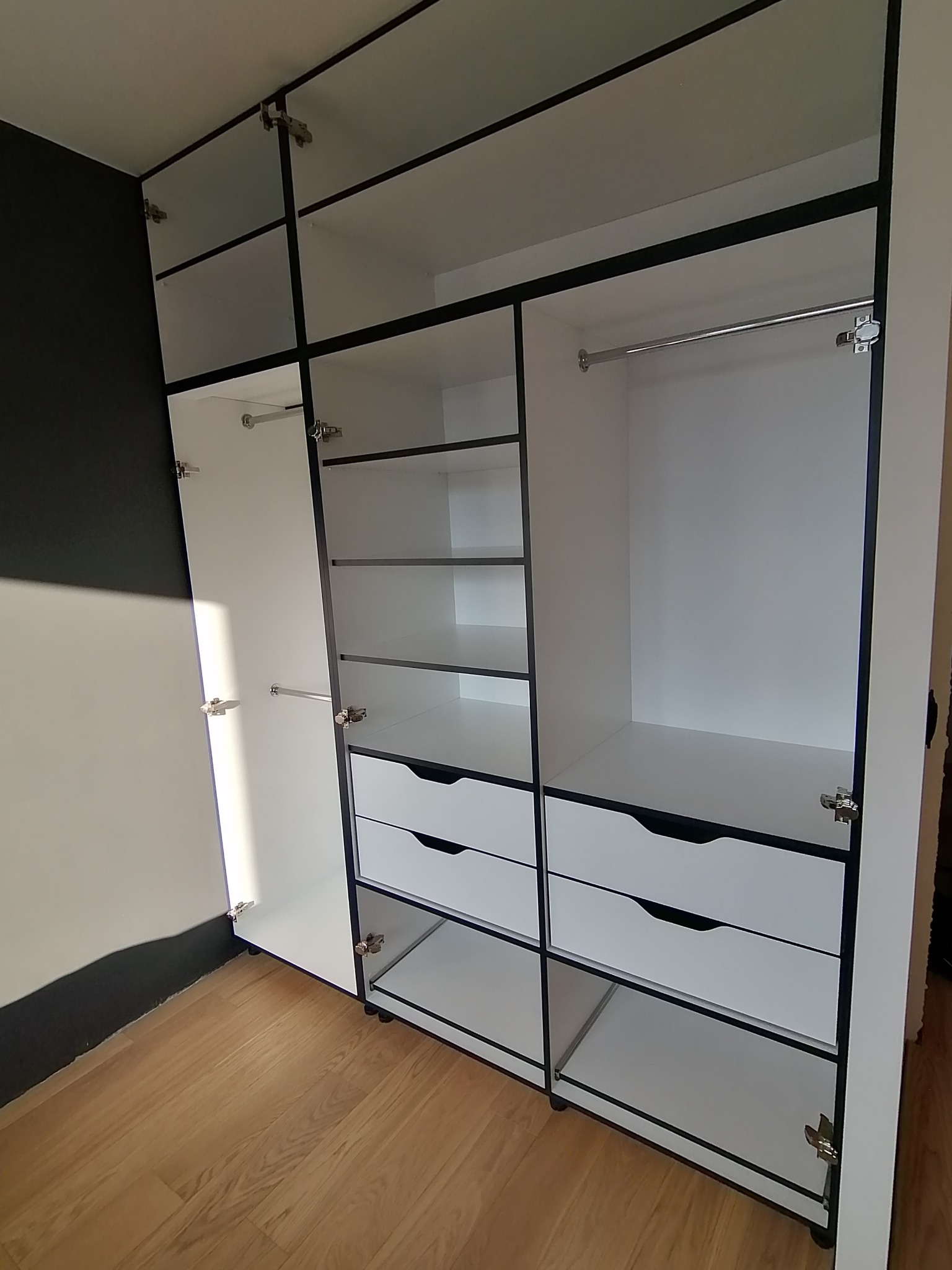 Biała szafa wnękowa z czarnymi obramowaniami, widoczne półki, szuflady i drążek na ubrania, jedne drzwi otwarte.