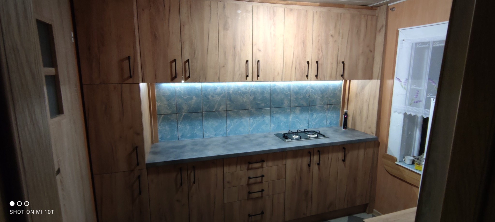 Kuchnia z drewnianymi szafkami na wymiar, szarym blatem i niebieskimi kaflami podświetlonymi LED, widoczna płyta gazowa.