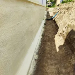 Świeżo nałożona, jasna izolacja na ścianie fundamentowej budynku, widoczna z poziomu wykopu, z niebieskim wężem w tle i domem jednorodzinnym w oddali.