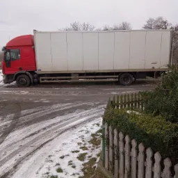 Czerwona ciężarówka z białą naczepą zaparkowana na błotnistym terenie z resztkami śniegu, widoczna zza drewnianego płotu.