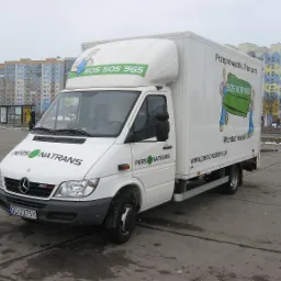 Biały samochód dostawczy marki Mercedes-Benz z logo 'PERSONATRANS' i numerem telefonu 505 505 965 na karoserii, zaparkowany na placu z blokami mieszkalnymi w tle.